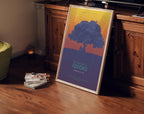 Un poster incorniciato di My Neighbor Totoro è appoggiato a un mobile di legno. Mostra un grande albero su sfondo sfumato arancione-blu, con Totoro sotto. Il titolo è in verde. Accanto, libri impilati e una candela accesa creano un’atmosfera calda.
